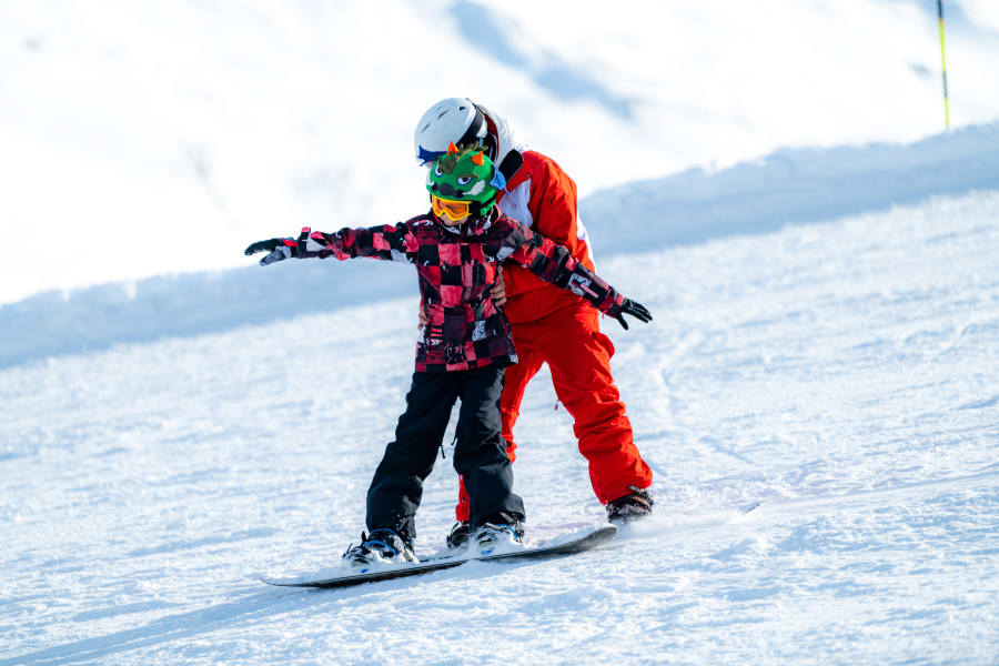 Child learning to snowboard with an adult holding on to them.